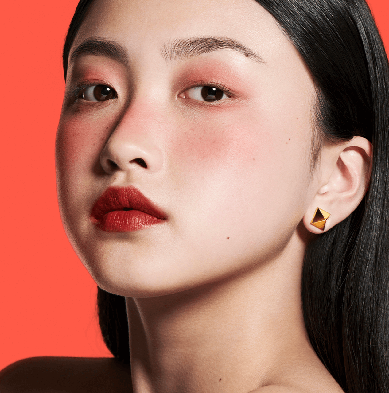 A close-up portrait of a woman with long black hair, wearing bold red lipstick and subtle eye makeup. She has a geometric earring and a smooth, even complexion against a bright coral background.