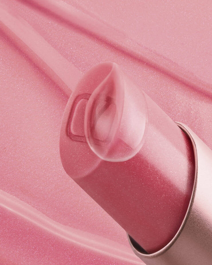 Un gros plan d'un baume a levres rose, avec son capuchon transparent et translucide legerement ouvert, revelant un subtil logo "V" a l'interieur, le tout sur un fond rose doux avec de legeres traces de produit.