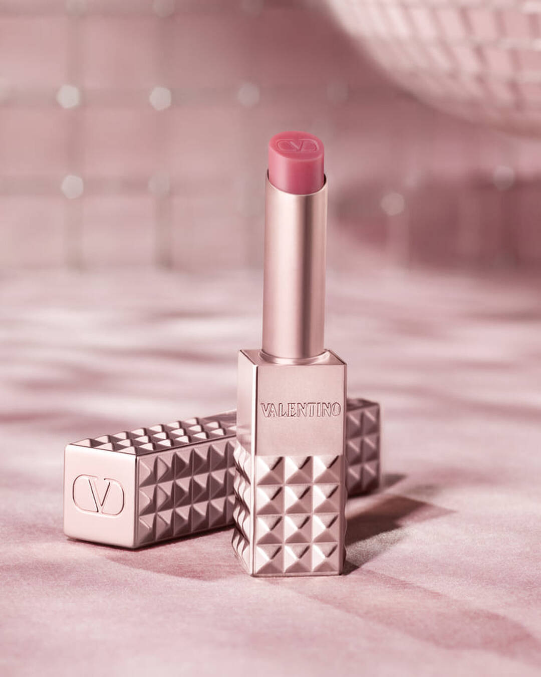 Un produit pour les levres Valentino Spike dore rose avec une pointe rose est presente debout, avec son etui cloute assorti pose a cote. Les deux articles sont positionnes sur un fond rose doux et scintillant qui evoque une sensation de glamour.